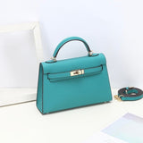 De Aurora Signature Mini Leren Handtas van Liora The Label, een turquoise gestructureerde handtas met gouden hardware en een bijpassende afneembare riem, rust op een wit oppervlak naast een wit fotolijstje en pastelkleurig papier.