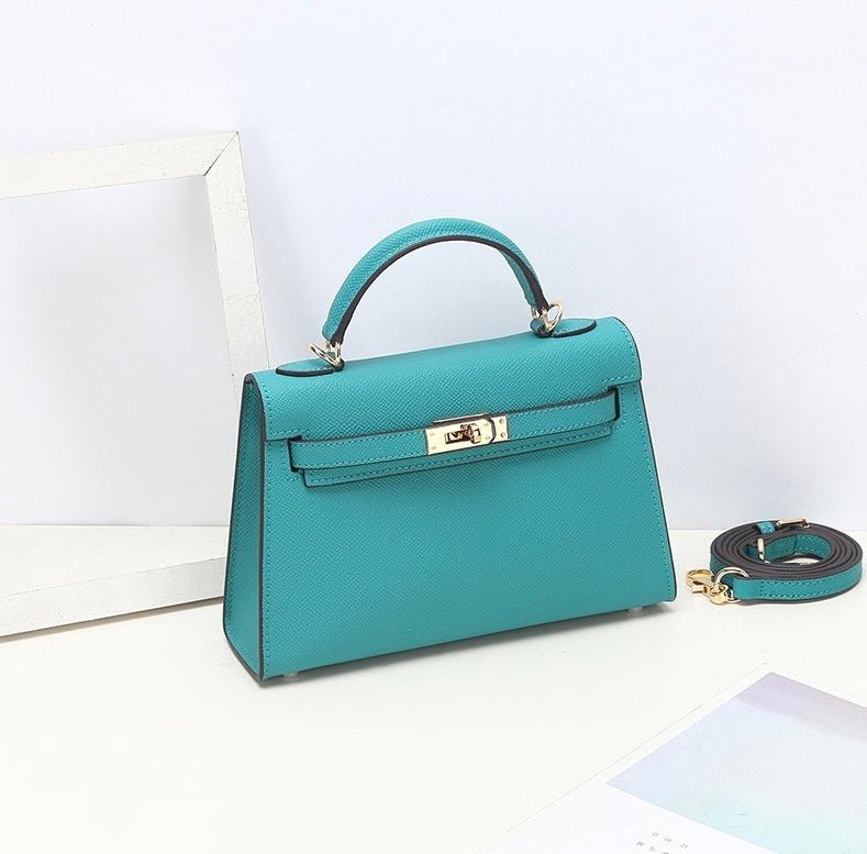 De Aurora Signature Mini Leren Handtas van Liora The Label, een turquoise gestructureerde handtas met gouden hardware en een bijpassende afneembare riem, rust op een wit oppervlak naast een wit fotolijstje en pastelkleurig papier.
