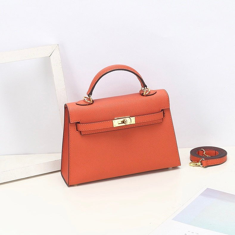 De Aurora Signature Mini Leren Handtas van Liora The Label is een oranje gestructureerde handtas met bovengreep in volnerf leer met gouden hardware, afgebeeld op een wit vlak naast de bijpassende afneembare schouderriem.