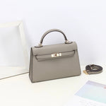 De Aurora Signature Mini Leren Handtas van Liora The Label in taupe volnerf leer heeft een handvat aan de bovenkant, gouden hardware en een sluiting aan de voorkant op een wit oppervlak, met een verstelbare riem vlakbij temidden van een wit frame en pastel papier.