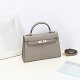 De Aurora Signature Mini Leren Handtas van Liora The Label in taupe volnerf leer heeft een handvat aan de bovenkant, gouden hardware en een sluiting aan de voorkant op een wit oppervlak, met een verstelbare riem vlakbij temidden van een wit frame en pastel papier.