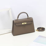 De Aurora Signature Mini Leren Handtas van Liora The Label, een taupe gestructureerde handtas met bovengreep en goudkleurige hardware en afneembare schouderriem, wordt getoond op een wit oppervlak naast een wit frame en pastelkleurig papier.