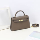 De Aurora Signature Mini Leren Handtas van Liora The Label, een taupe gestructureerde handtas met bovengreep en goudkleurige hardware en afneembare schouderriem, wordt getoond op een wit oppervlak naast een wit frame en pastelkleurig papier.