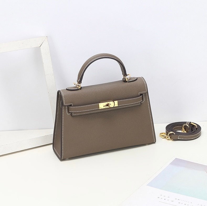 De Aurora Signature Mini Leren Handtas van Liora The Label, een taupe gestructureerde handtas met bovengreep en goudkleurige hardware en afneembare schouderriem, wordt getoond op een wit oppervlak naast een wit frame en pastelkleurig papier.