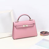 De Aurora Signature Mini Leren Handtas van Liora The Label is een roze, gestructureerde tas met bovenhandvat in volnerf leer met gouden hardware en gespsluiting. De tas heeft een afneembare riem en rust op een wit vlak bij een wit fotolijstje.