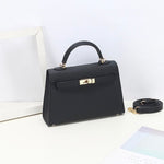 De Aurora Signature Mini Leren Handtas van Liora The Label - een zwarte, gestructureerde tas met bovenhandgreep, gouden hardware en een afneembare schouderriem - rust op een wit oppervlak naast een lege lijst en een pastelkleurig blad.