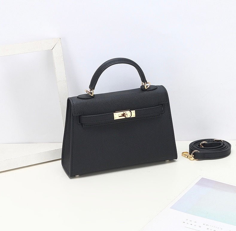 De Aurora Signature Mini Leren Handtas van Liora The Label - een zwarte, gestructureerde tas met bovenhandgreep, gouden hardware en een afneembare schouderriem - rust op een wit oppervlak naast een lege lijst en een pastelkleurig blad.