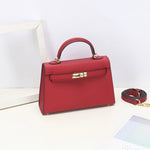 De Aurora Signature Mini Leren Handtas van Liora The Label is een gestructureerde top handtas van volnerf leer met een gouden sluiting en afneembare schouderriem, gepresenteerd op een witte ondergrond naast een wit frame en deels zichtbaar papier.