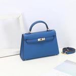 De Aurora Signature Mini Leren Handtas van Liora The Label, een gestructureerde blauwe minitas in volnerf leer met een bovenhandvat en gouden hardware, staat op een wit vlak met de bijpassende schouderriem ernaast.