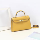 De Aurora Signature Mini Leren Handtas van Liora The Label in geel volnerf leer, met top handvat, gouden sluiting en afneembare schouderriem, wordt op een witte ondergrond bij een lege witte fotolijst getoond.