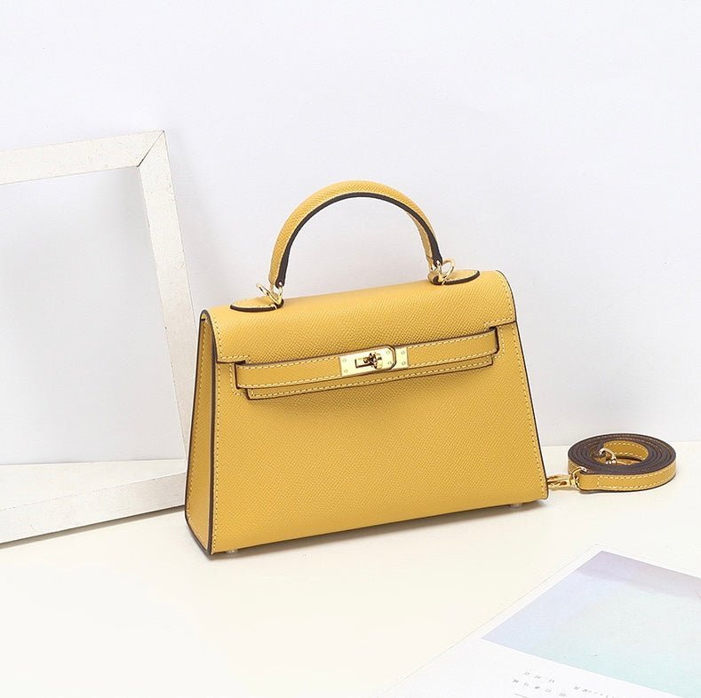 De Aurora Signature Mini Leren Handtas van Liora The Label in geel volnerf leer, met top handvat, gouden sluiting en afneembare schouderriem, wordt op een witte ondergrond bij een lege witte fotolijst getoond.