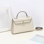 De Aurora Signature Mini Leren Handtas van Liora The Label, in crème volnerf leer met een bovenhandgreep, gouden hardware en een afneembare schouderriem, wordt tentoongesteld op een wit oppervlak naast een fotolijstje en een tijdschrift.