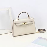 De Aurora Signature Mini Leren Handtas van Liora The Label, in crème volnerf leer met een bovenhandgreep, gouden hardware en een afneembare schouderriem, wordt tentoongesteld op een wit oppervlak naast een fotolijstje en een tijdschrift.