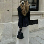 Een vrouw met lang, golvend blond haar staat op een stenen stoep, met haar gezicht naar voren, en houdt de Elaris Luxe Taupe Leren Emmer Tas van Liora The Label vast - een elegante, tijdloze tas - in een stedelijke omgeving. Ze draagt een sjaal met patroon en een zwarte blazer.