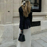 Een vrouw met lang, golvend blond haar staat op een stenen stoep, met haar gezicht naar voren, en houdt de Elaris Luxe Taupe Leren Emmer Tas van Liora The Label vast - een elegante, tijdloze tas - in een stedelijke omgeving. Ze draagt een sjaal met patroon en een zwarte blazer.