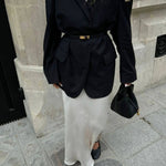 Een persoon draagt een zwarte oversized blazer met riem, een lange witte satijnen rok, zwarte flats en houdt de Elaris Luxe Taupe Leren Emmer Tas van Liora The Label vast, staand op een stoep in de stad bij een stenen gebouw.