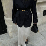 Een persoon draagt een zwarte oversized blazer met riem, een lange witte satijnen rok, zwarte flats en houdt de Elaris Luxe Taupe Leren Emmer Tas van Liora The Label vast, staand op een stoep in de stad bij een stenen gebouw.