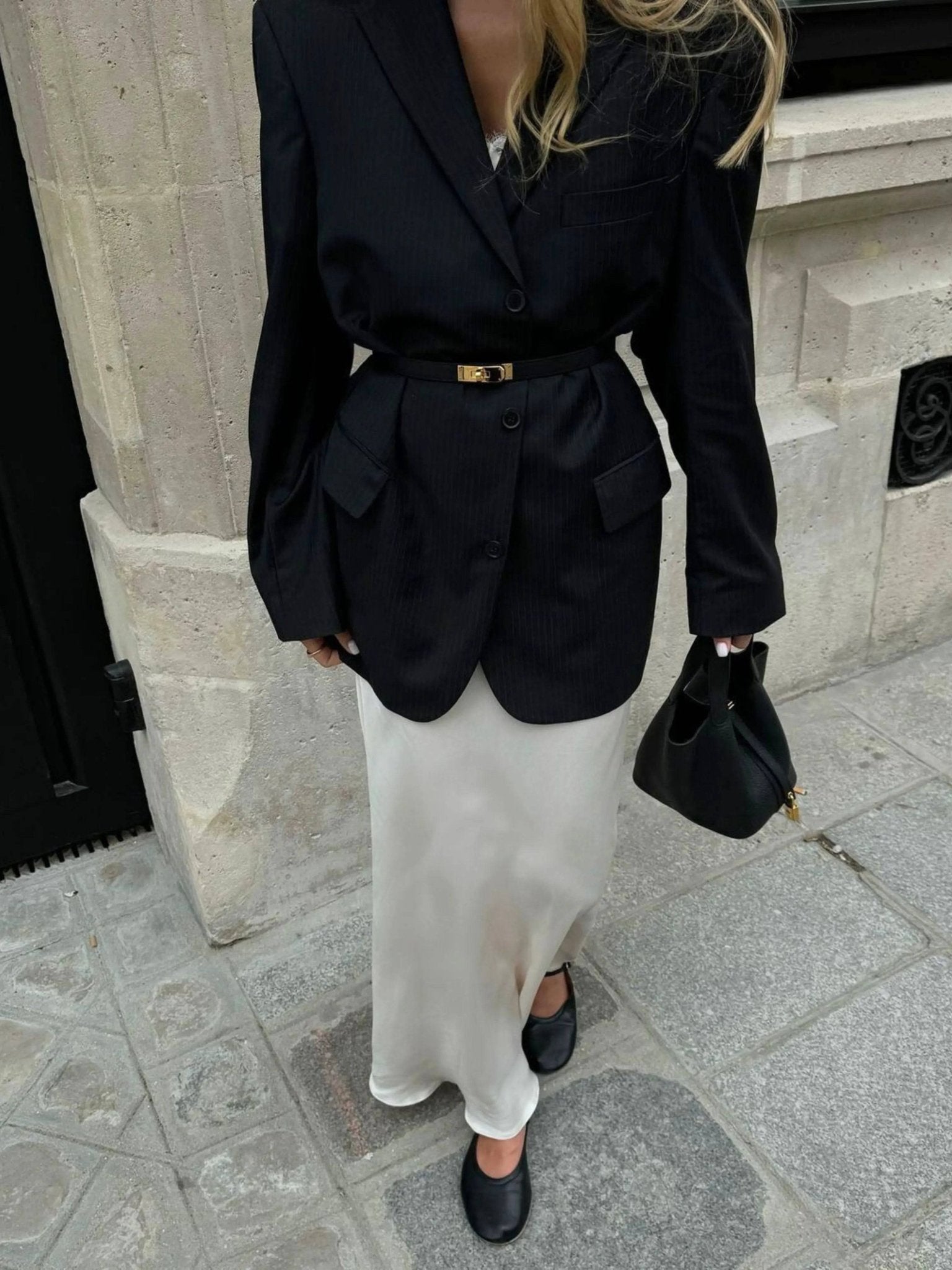 Een persoon draagt een zwarte oversized blazer met riem, een lange witte satijnen rok, zwarte flats en houdt de Elaris Luxe Taupe Leren Emmer Tas van Liora The Label vast, staand op een stoep in de stad bij een stenen gebouw.