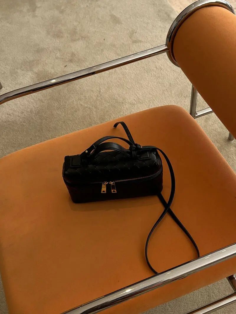 De Luxe Geweven Box Bag van Liora The Label, een elegante donkerbruine crossbody met gouden ritssluiting, rust op de zitting van een oranje stoel met kussens en chromen armleuningen op een lichtgekleurd tapijt.