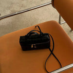 De Luxe Geweven Box Bag van Liora The Label, een elegante donkerbruine crossbody met gouden ritssluiting, rust op de zitting van een oranje stoel met kussens en chromen armleuningen op een lichtgekleurd tapijt.