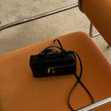 De Luxe Geweven Box Bag van Liora The Label, een elegante donkerbruine crossbody met gouden ritssluiting, rust op de zitting van een oranje stoel met kussens en chromen armleuningen op een lichtgekleurd tapijt.