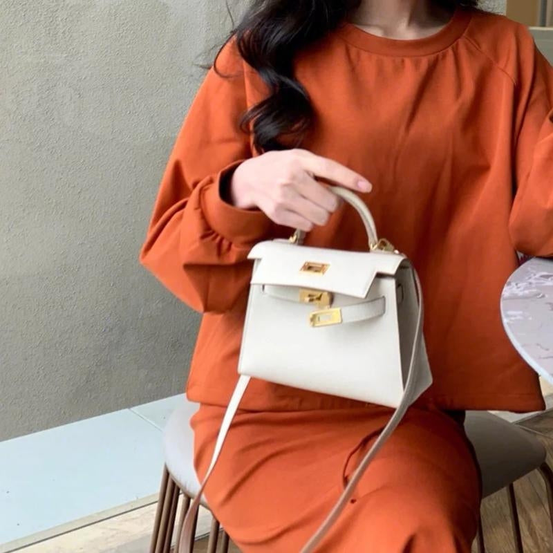 Een vrouw in een oranje jurk zit op een stoel en houdt de Galatea Elegante Zwarte Leren Handtas van Liora The Label vast, gemaakt van veganistisch saffiano-leer met gouden beslag. Haar gezicht is gedeeltelijk buiten beeld, donker golvend haar valt als ze bij een ronde tafel zit.